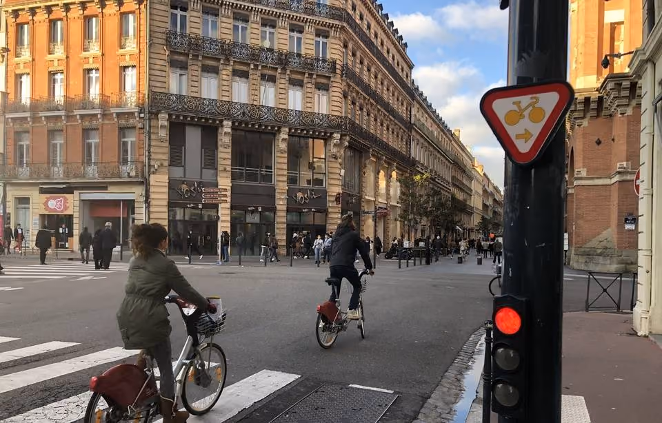 Photo d’un carrefour. Sur le côté droit, un feu tricolore avec au-dessus un tout petit panneau triangulaire inversé avec dedans un vélo jaune et une flèche de direction.